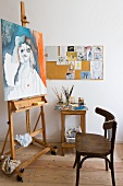 Simple wooden chair in front of painting on easel and painting utensils on side table against wall