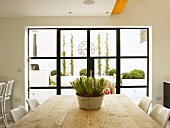 Blumentopf auf rustikalem Holztisch vor schwarzem Sprossenfenster und Blick in modern gestalteten Innenhof