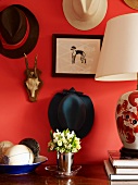 Posy in metal beaker flanked by table lamp and dish of ornamental fruit below collection of hats hanging on red-painted wall