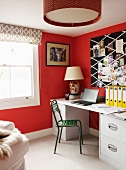 Green metal chair in front of pale desk against red-painted wall and patterned blind on window