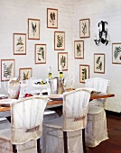 Stühle mit Husse an gedecktem Esstisch in Zimmerecke mit gerahmten Farnmotiven an weiß getünchter Ziegelwand.