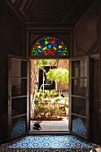 Inviting view of sunny courtyard planted with small trees and bushes from dark Oriental interior through open French doors with colourful stained glass in fanlight