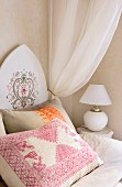 Embroidered cushion on bed with Oriental-style painted headboard and white, gathered canopy; spherical, white table lamp with simple lampshade on bedside cabinet