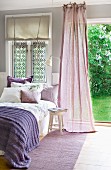 Comfortable bed with purple throw next to fluttering, mauve curtain in front of open door leading to garden