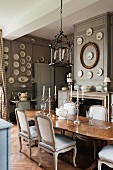 Candlesticks on wooden table and pale, upholstered chairs in grand dining room with wall plates on grey-painted, wood-panelled walls