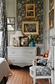 Corner of traditional bedroom with chest of drawers painted pale grey and gilt-framed pictures on wall with wallpapered inserts