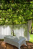 Secluded area in garden - daybed with white covers on terracotta tiled floor and curtains below tree canopy with spider web of struts