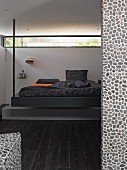 Designerbett mit Metallstange von der Decke abgehängt vor umlaufendem Fensterband, im Vordergrund mit Kieselmosaik geflieste Wand und dunkler Parkettboden