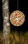 Spherical, perforated, orange pendant lamp hanging next to birch trunk against dark, night-time background like a lantern