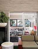 Upholstered sofa on terrace against house facade with view into bright dining room with plexiglass furniture