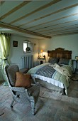 Einfaches Schlafzimmer mit Terrakottafliesen und niedriger Balkendecke; in der Mitte ein gemütliches Doppelbett mit rustikalem Kopfende aus Holz