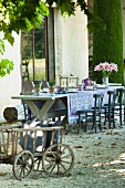 Dining table set with blue tablecloth and flowers on gravel terrace outside French country-house; cushions in pull-along wagon in foreground