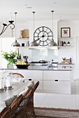 White, open-plan kitchen with pendant lamps, wall clock, rustic dining table and wicker chairs