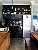 Corner of simple, black kitchen with stainless steel fridge-freezer and partially visible wooden table and bench in traditional setting