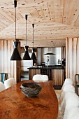Chalet dining area with polished, live-edge, wooden table top, black designer lamps and view into kitchen area