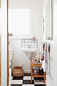 Einblick in kleines Badezimmer mit Holzregal und Waschbecken an weißer Wand, am Boden schwarz-weiß gefliestes Schachbrettmuster