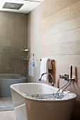 Freistehende Badewanne in modernem Badezimmer mit glänzender Wasserfallarmatur und Badewannenablage; dahinter der Duschbereich mit Glastrennwand