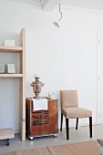 Silver samovar on vintage tea chest between upholstered chair and wooden shelves