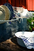 Washing-up bowl on wooden table below drying rack of crockery hung on house facade