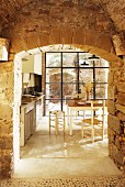 Blick auf einfachen Essplatz in lichtdurchfluteter Küche durch den bogenförmigen Durchgang in alter Natursteinmauer