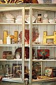 Vitrinenschrank gefüllt mit einem Sammelsurium an Vintage- Objekten, von Geschirr bis zum Bilderrahmen