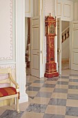 Antique grandfather clock with gilt embellishments against wall between two open doors and chequered floor in foyer of stately home