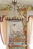 Kronleuchter mit kleinen weissen Schirmen vor bemalter Wand und Kerzenständer auf goldverziertem Konsolentisch