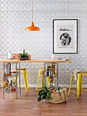 Kitchen table with wooden top and storage below on metal shelving; metal stools and industrial-style, retro pendant lamp in front of embossed metal wall panels