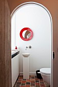 Weisser Toilettenraum mit kleinem Standwaschbecken und Terrakottafliesen; darüber ein rotgerahmter, runder Spiegel