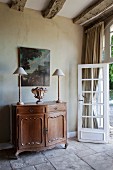 Two table lamps on Rococo cabinet below oil painting on wall next to open French doors