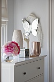 Flowers, vases, box and female hand as jewellery stand on white chest of drawers