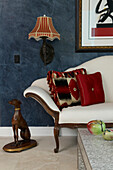 Detail of upholstered bench with scatter cushions leaning on arm next to animal figurine against blue marbled wall with traditional sconce lamp