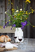 Cheese, bread, two glasses of red wine and a milk churn of wild flowers on a rustic wooden table