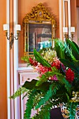 Tropical bouquet on table in front of apricot wall with gilt-framed mirror flanked by sconce lamps