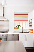 View over a table to a kitchen sink at a window, with a half-closed Roman blind with colorful stripes next to a gas oven in a modern kitchen