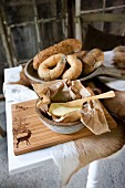 Wooden knife in butter dish on wooden chopping board in front of bread rolls in ceramic dish on table covered with animal skin