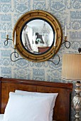 Round framed mirror on wallpapered wall with candle holders on the bed with wooden headboard
