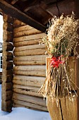 Sheaf of cereal tied to wooden structure with red ribbon in front of outer wall of log cabin