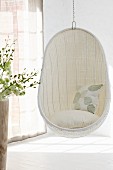 Comfortable, white wicker hanging chair in corner of room next to window with closed, translucent curtains and pattern of light and shadow on floor