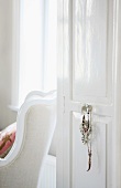 Open, white-painted interior door with costume jewellery hanging from doorknob and view of traditional armchair beyond