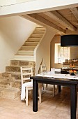 Black wooden table and simple kitchen chairs in front of stone and wood staircase in open-plan, rustic interior with wood-beamed ceiling