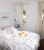 Straw hat on double bed with striped bed linen next to window with floor-length curtains in white bedroom