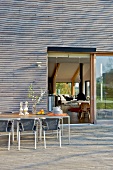 Table and chairs on terrace of contemporary house with exterior wood cladding and view of living room through open terrace door