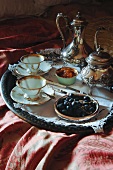 Frühstückstablett auf dem Bett mit silbernen Rokokokannen, goldverzierten Porzellantassen und Weintrauben