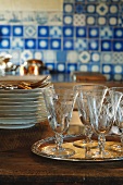 Old, etched wine glasses on silver tray in front of stacked plates; Delft blue antique tiles blurred in background