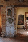 Dekorativ gestaltete, alte Holztür vor Ziegelwand im rustikalen Hausflur eines französischen Landhauses; historisches Ölgemälde im Hintergrund