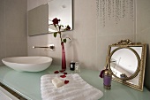 Badezimmerutensilien auf Designerwaschtisch mit Waschschüssel auf Glasplatte und Blumenvase neben Vintage Spiegel