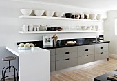 Bowls on white floating shelves above kitchen counter with grey units in open-plan, modern, designer kitchen