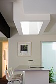 Shaft-shaped skylight above a kitchen island clad with stainless steel and a row of white bar stools
