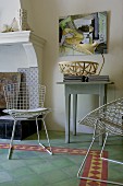 White, wire mesh chairs and small, rustic console table on green tiled floor with border next to open fireplace in living room